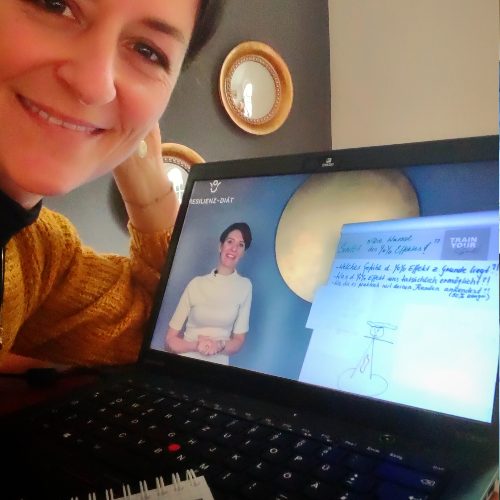 Frau neben Laptop, auf dessen Bildschirm ein Video mit einer Dozentin zu sehen ist.