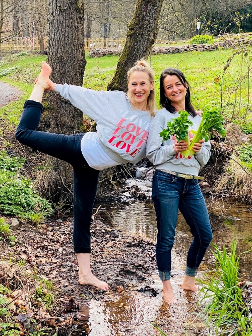 Zwei Frauen barfuß im Bach. Die eine Macht die Yogapose Tänzer, die andere hält zwei Staudensellerie in den Händen.