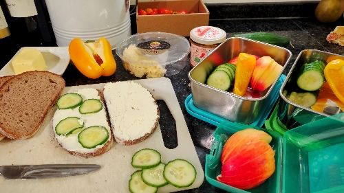 Brotboxen zum Teil gefüllt mit Apfel, Gurken und Paprika. Auf einem Küchenbrett Brotscheiben, die mit Frischkäse geschmiert und mit Gurkenscheiben belegt werden.